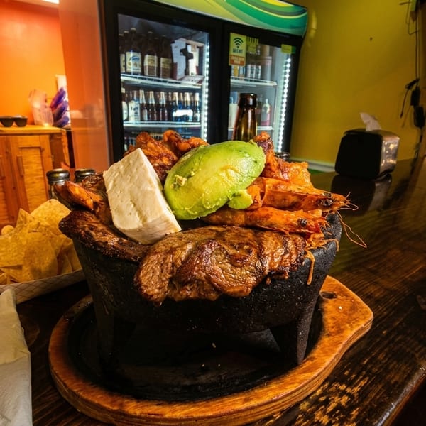 Hearty molcajete with grilled steak, shrimp, avocado, and cheese on a bar, with chips and beer menu.