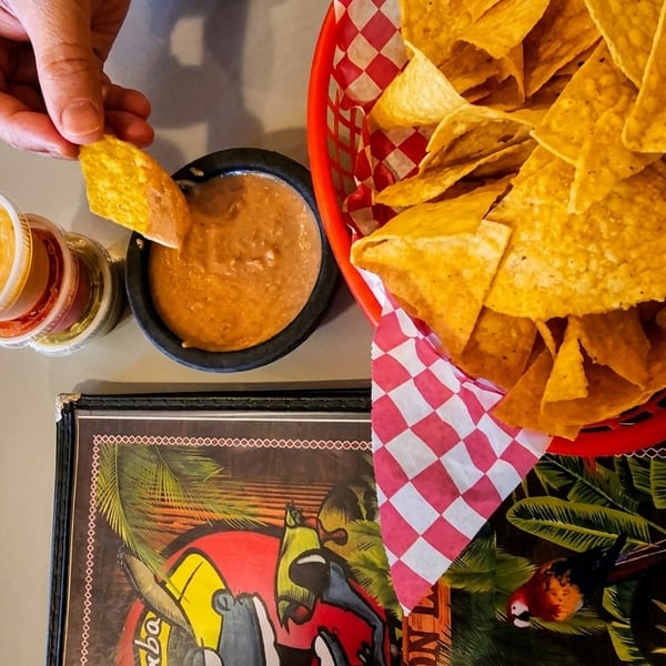 Hand dipping a tortilla chip into a bowl of bean dip next to a basket of chips and small sauces.