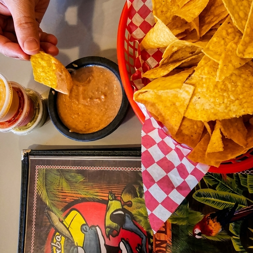 Hand dipping a tortilla chip into a bowl of bean dip next to a basket of chips and small sauces.