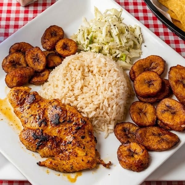 Grilled chicken, rice, fried plantains, and coleslaw on a white plate with a checkered tablecloth.