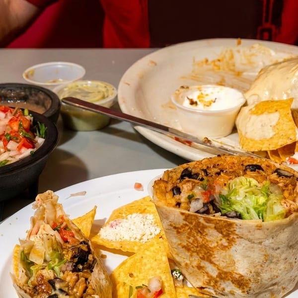 Close-up of two plates with large burritos, tortilla chips, and side dips on a table.
